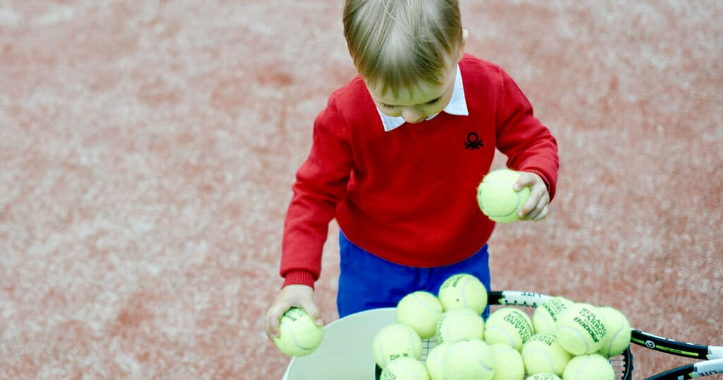 Kid on a Red Artificial Grass Tennis Court Collects Tennis Balls into the Bucket